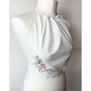 May include: White cotton halter top with a scalloped edge and a floral embroidery design. The top has a tie closure at the back.