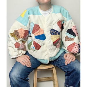 May include: A white quilted jacket with a blue collar and a patchwork design featuring colorful pinwheels. The jacket is worn over a white t-shirt and blue jeans.