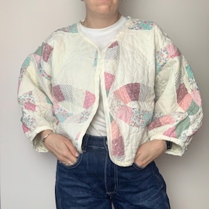 May include: A white quilted jacket with a patchwork design featuring pink, green, and blue floral patterns. The jacket has a button-up front and long sleeves.