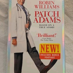 May include: A sealed VHS tape of the movie "Patch Adams" starring Robin Williams. The cover features Williams in a doctor's coat, with the film's title in large red letters. A "NEW!" sticker and the words "Includes Bonus Footage" are also visible.