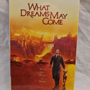 May include: A VHS video cassette cover for the movie "What Dreams May Come" starring Robin Williams, Cuba Gooding Jr., and Annabella Sciorra. The cover features a cityscape with a man walking with a dog in the foreground. The text "After Life There Is More" is printed on the cover.