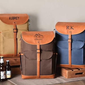 Puede incluir: Tres mochilas de lona y cuero con nombres personalizados. Las mochilas son marr&oacute;n, beige y azul. Los nombres en las mochilas son Aubrey, Vincent y RK.