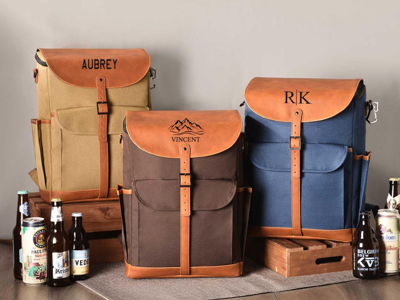 Puede incluir: Tres mochilas de lona y cuero con nombres personalizados. Las mochilas son marr&oacute;n, beige y azul. Los nombres en las mochilas son Aubrey, Vincent y RK.