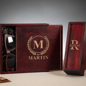 Op de afbeelding: Twee houten dozen met gegraveerde initialen en namen. De grotere doos is bordeauxrood met een gouden lauwerkrans en de initialen "M" erin. De tekst "MARTIN" is onder de krans gegraveerd. De kleinere doos is bordeauxrood met de initialen "R" en de tekst "ROBERT" gegraveerd op de voorkant.