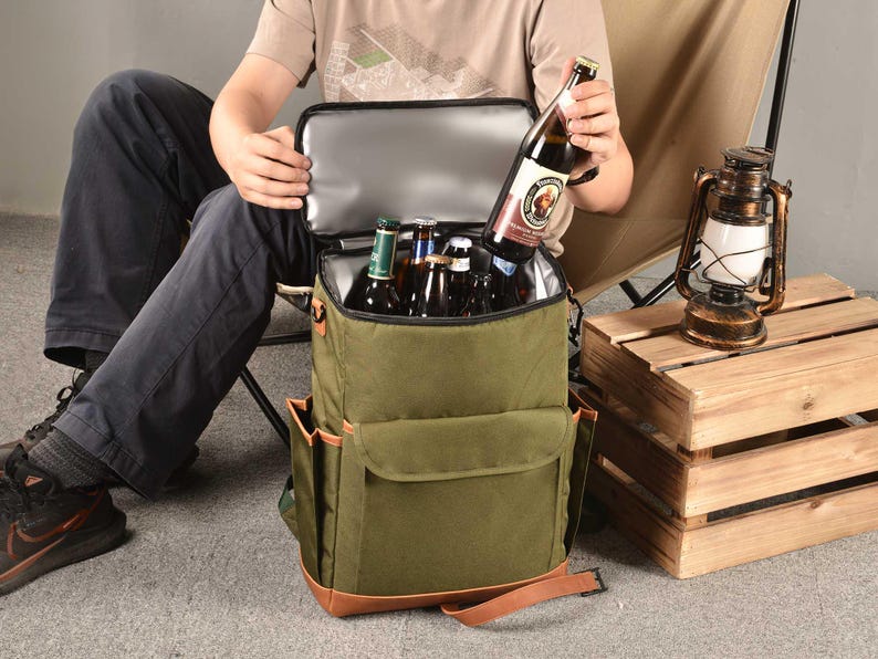 Puede incluir: Una mochila enfriadora de lona verde con detalles en cuero marr&oacute;n y un bolsillo frontal con cremallera. El enfriador est&aacute; lleno de seis botellas de cerveza.