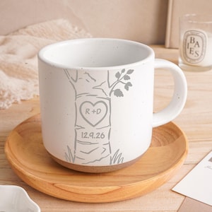 Puede incluir: Una taza de cerámica blanca con acabado moteado y un gráfico gris de un tronco de árbol. El árbol presenta un grabado en forma de corazón con las letras R+D y la fecha 12.9.26 debajo. La taza tiene un asa y una base de color marrón.
