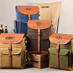 Puede incluir: Cuatro mochilas de lona personalizadas con detalles de cuero. Las mochilas son de diferentes colores, incluyendo marr&oacute;n, verde, azul y beige. Cada mochila tiene un nombre diferente en la parte delantera, incluyendo "Aubrey", "Vincent", "RTS" y "RK".