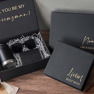 May include: A set of black gift boxes with gold lettering. One open box contains a black tumbler, black sunglasses, and a black wallet nestled in silver paper filler. The inside lid reads WILL YOU BE MY Groomsman? Two closed boxes are labeled Noah GROOMSMAN and Liam BEST MAN.