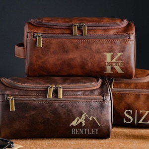 Könnte beinhalten: Drei braune Kulturbeutel aus Leder mit goldenen Reißverschlüssen und personalisiertem Text. Eine Tasche trägt den Namen "Kenneth", eine andere "SIZ" und die dritte "Bentley" mit einem Bergmotiv. Die Taschen sind übereinander gestapelt.