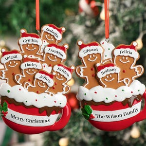 Könnte beinhalten: Zwei Weihnachtsschmuckstücke in Form von Lebkuchenmännchen in einer rot-weißen Tasse. Auf einem steht "Merry Christmas" und auf dem anderen "The Wilson Family". Jedes Lebkuchenmännchen hat einen Namen auf seinem Hut.