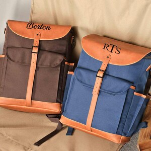 Puede incluir: Dos mochilas de lona personalizadas con detalles de cuero. La mochila de la izquierda es marr&oacute;n con el nombre "Berton" en la solapa. La mochila de la derecha es azul con las iniciales "RTS" en la solapa.
