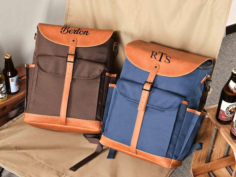 Puede incluir: Dos mochilas de lona personalizadas con detalles de cuero. La mochila de la izquierda es marr&oacute;n con el nombre "Berton" en la solapa. La mochila de la derecha es azul con las iniciales "RTS" en la solapa.