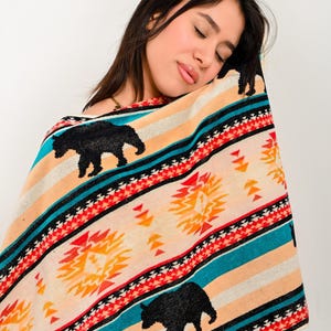 Peut inclure: Une couverture colorée avec un motif amérindien représentant des ours noirs, des motifs géométriques et des rayures dans les tons orange, turquoise et crème. La couverture a une texture douce et peut être utilisée comme jeté ou enveloppe.