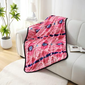 May include: A pink blanket with a geometric pattern in shades of pink, blue, and white, draped over a white sofa. The blanket has a black border. The sofa sits on a white rug.