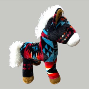 Pu&ograve; includere: Un giocattolo a forma di cavallo di peluche con un colorato motivo geometrico rosso, nero e turchese. Il giocattolo ha criniera e coda bianche e zoccoli marroni. Il cavallo &egrave; su sfondo grigio.