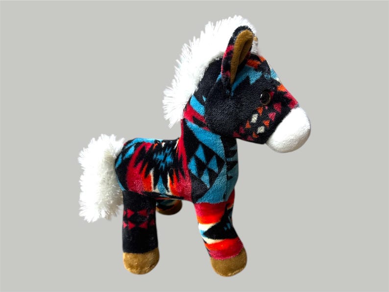 Pu&ograve; includere: Un giocattolo a forma di cavallo di peluche con un colorato motivo geometrico rosso, nero e turchese. Il giocattolo ha criniera e coda bianche e zoccoli marroni. Il cavallo &egrave; su sfondo grigio.