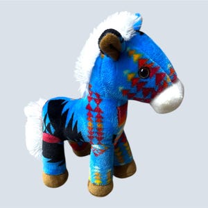 Pu&ograve; includere: Un cavallo di peluche blu con un colorato motivo geometrico rosso, giallo e nero. Ha criniera e coda bianche, orecchie marroni e muso bianco. Il cavallo &egrave; alto circa 20 cm.