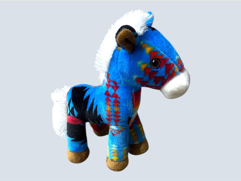 Pu&ograve; includere: Un cavallo di peluche blu con un colorato motivo geometrico rosso, giallo e nero. Ha criniera e coda bianche, orecchie marroni e muso bianco. Il cavallo &egrave; alto circa 20 cm.