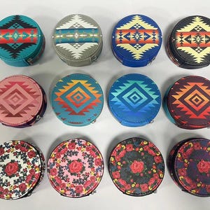 Pode incluir: Uma coleção de estojos redondos para joias com zíper. Os estojos apresentam vários designs, incluindo padrões geométricos em turquesa, laranja e preto, e padrões florais com rosas vermelhas. Os estojos são exibidos em uma superfície branca.