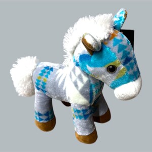 Pu&ograve; includere: Un cavallo di peluche con criniera e coda bianche. Il corpo del cavallo &egrave; grigio chiaro con un motivo geometrico blu e giallo. Gli zoccoli sono marroni e le orecchie blu e marroni.