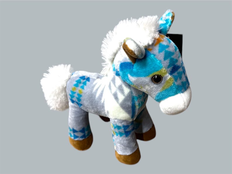 Pu&ograve; includere: Un cavallo di peluche con criniera e coda bianche. Il corpo del cavallo &egrave; grigio chiaro con un motivo geometrico blu e giallo. Gli zoccoli sono marroni e le orecchie blu e marroni.
