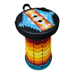 May include: A colorful, collapsible stool with a black top featuring a geometric design in turquoise, orange, and white. The accordion-style body is made of red, orange, yellow, and blue plastic. A black handle is attached to the top.