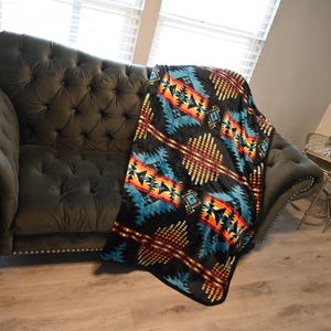 May include: A black blanket with a Southwestern geometric pattern in shades of turquoise, orange, and yellow is draped over a dark gray tufted sofa. The blanket's design features diamond and arrow shapes.