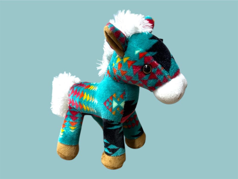 Pu&ograve; includere: Un cavallo di peluche con un corpo turchese con un colorato motivo geometrico in rosso, giallo e arancione. Il cavallo ha criniera e coda bianche, zoccoli e orecchie marroni. Il giocattolo &egrave; su uno sfondo azzurro.