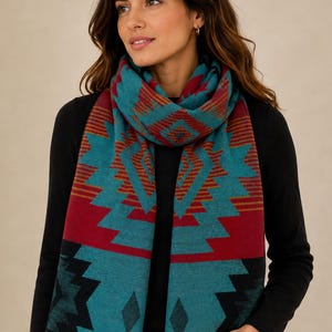 Puede incluir: Una bufanda rectangular con un patrón geométrico en color verde azulado, rojo intenso y negro. El diseño incluye formas de diamante y escalonadas repetidas con bandas de rayas horizontales. La bufanda tiene un borde con flecos.