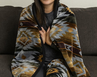 May include: A person wrapped in a soft, patterned blanket. The blanket features a geometric design in shades of gray, gold, brown, and black. The blanket is draped over the person, who is sitting on a dark gray couch.