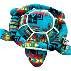 May include: A plush turtle toy in shades of turquoise, red, yellow, and black. The turtle's shell and limbs feature a Southwestern-inspired geometric pattern. The toy has a white trim and a tag attached.