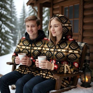 Könnte beinhalten: Zwei Personen tragen passende Kapuzenjacken mit einem geometrischen Muster in Braun, Schwarz, Rot und Beige. Sie sitzen auf einer Holzschaukel und halten weiße Tassen, mit einem verschneiten Wald im Hintergrund. Die Jacken haben einen Reißverschluss.