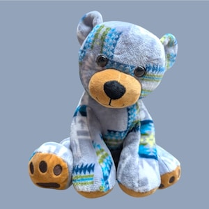 May include: A plush teddy bear with a light gray body and patterned patches of blue, green, and white. The bear has a tan snout, brown paws, and black eyes. The bear is sitting with its legs extended.