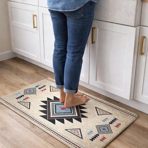 Puede incluir: Una alfombra de cocina rectangular beige con un patrón geométrico en negro, rojo y azul. El diseño incluye triángulos, diamantes y un motivo cuadrado central. La alfombra está colocada sobre un suelo de madera.