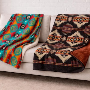 May include: Two patterned throw blankets rest on a beige sofa. One blanket features a turquoise, red, and orange geometric design. The other blanket has a black, tan, and brown geometric pattern. The blankets are made of soft material.