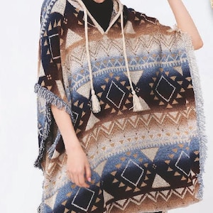 May include: A hooded poncho with a geometric pattern in shades of brown, blue, and cream. The poncho has fringe along the edges and a drawstring with tassels. A straw hat with a black band is also visible.