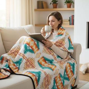 Op de afbeelding: Een witte deken met een geometrisch patroon in oranje en teal is over een beige bank gedrapeerd. De deken is om een persoon gewikkeld die een mok en een boek vasthoudt. Een hond rust op de houten vloer bij een open haard.
