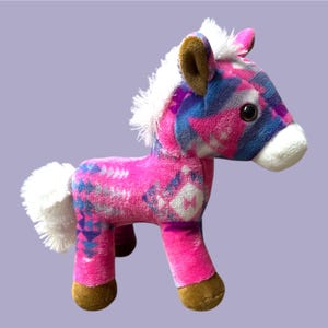 Pu&ograve; includere: Un cavallo di peluche rosa, blu e bianco. Ha criniera e coda bianche, zoccoli e orecchie marroni e grandi occhi neri. Il corpo presenta un motivo.