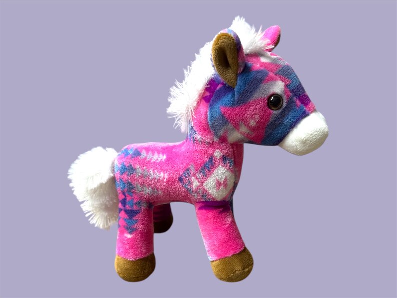 Pu&ograve; includere: Un cavallo di peluche rosa, blu e bianco. Ha criniera e coda bianche, zoccoli e orecchie marroni e grandi occhi neri. Il corpo presenta un motivo.