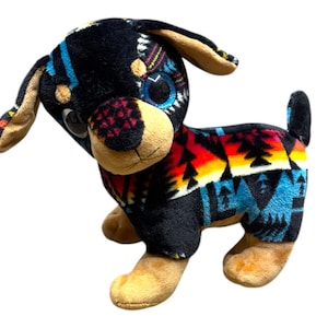 May include: A plush dog toy with a black body and tan accents. The dog's body is decorated with a colorful geometric pattern in shades of red, orange, yellow, and blue. The dog has large, expressive eyes.