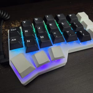 May include: Ergonomic split keyboard with black keycaps and white accents. The keys are backlit with blue and green LEDs. The keyboard has a curved design and a wired connection.