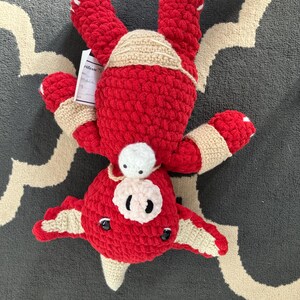 May include: A handmade red crocheted creature with beige detailing, including a hat, arm bands, and wing-like features. The plush toy has black button eyes and a white nose. A tag is attached to the toy.