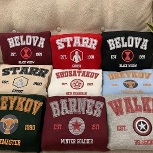 May include: Ten different colored sweatshirts with embroidered logos and text. The sweatshirts are arranged in three rows. The top row has three sweatshirts, the middle row has three sweatshirts, and the bottom row has four sweatshirts. The sweatshirts have different colors, including red, blue, green, gray, and beige. The text on the sweatshirts includes the names "BELOVA", "STARR", "SHOSATAKOV", "DREYKOV", "BARNES", and "WALKER".