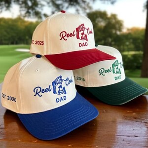 Puede incluir: Tres gorras de béisbol con coronas blancas y viseras de colores. Una tiene visera roja y bordado rojo que dice Reel Cool DAD EST. 2025. Una tiene visera azul y bordado azul Reel Cool DAD EST. 2020. Una tiene visera verde y bordado verde.