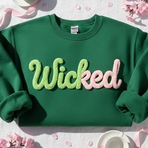 Puede incluir: Una sudadera verde oscuro con la palabra "Wicked" en una fuente texturizada verde claro y rosa. La sudadera tiene cuello redondo y mangas largas. El fondo incluye flores rosas, una cinta y una taza de té.