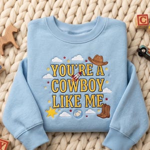 May include: A light blue crewneck sweatshirt featuring yellow embroidered text that reads YOU'RE A COWBOY LIKE ME. The design includes embroidered icons of a brown cowboy hat, a rocket ship, a pair of brown boots, a yellow star, and white clouds.