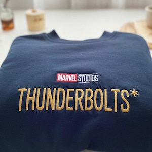 Peut inclure: Un sweat-shirt bleu marine avec un logo "THUNDERBOLTS*" brodé en or et un logo "MARVEL STUDIOS" rouge.