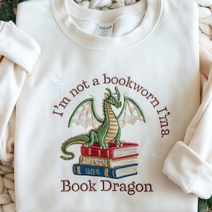 Op de afbeelding: Crèmekleurige sweatshirt met geborduurd ontwerp. Het ontwerp toont een groene draak bovenop een stapel boeken. De tekst luidt "I'm not a bookworm I'm a. Book Dragon" in bruine draad. De mouwen zijn opgerold.