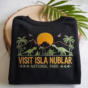 以下が含まれることがあります： 黄色い太陽、ヤシの木、山、2匹の緑色の恐竜が刺繍されたデザインの黒いスウェットシャツ。「VISIT ISLA NUBLAR NATIONAL PARK」と書かれています。
