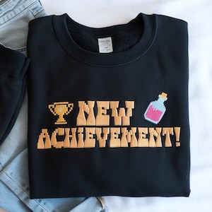 Może przedstawiać: Czarna bluza z haftowanym wzorem w kolorze złotym, na którym widnieje napis "NEW ACHIEVEMENT!" wraz z pikselowanym trofeum i miksturą.
